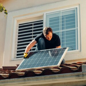 A worker installing a solar panel on a residential rooftop under bright sunlight.